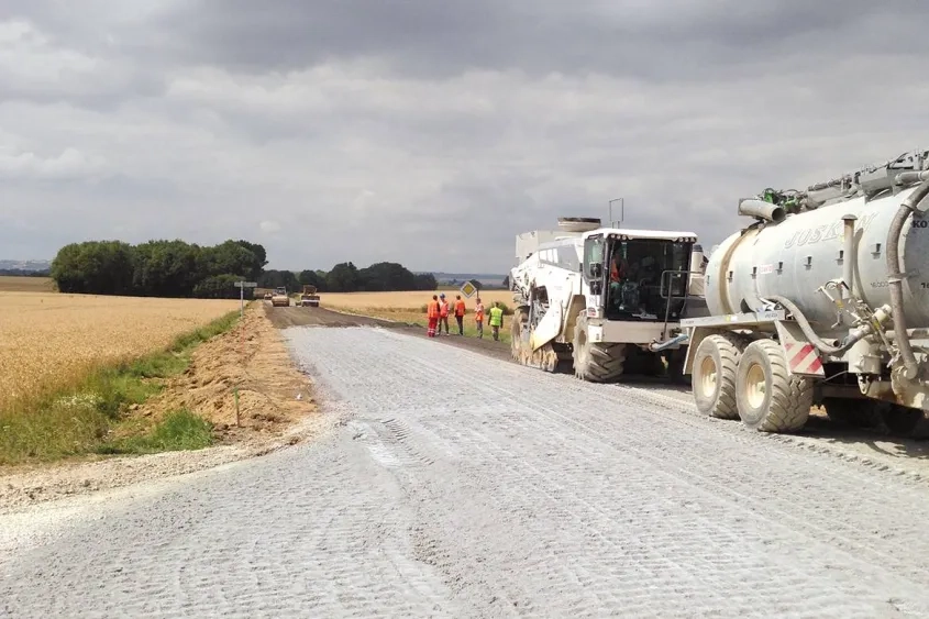 Vue générale du chantier avec, au premier plan, la machine de malaxage alimentée en eau par une citerne, et en arrière plan, la niveleuse et l'atelier de compactage.