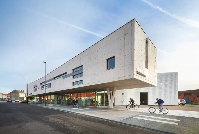 L’entrée vitrée et le parvis de la médiathèque se glissent sous le volume suspendu : parallélépipède rectangle en béton brut clair qui contraste avec les maisons environnantes.